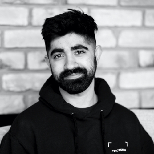 Manav Kher is smiling at the camera with a brick wall behind him.